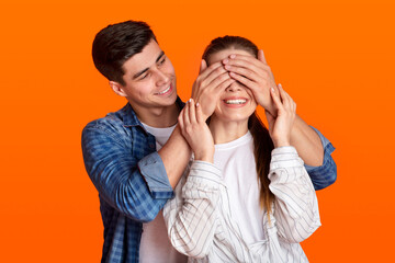 Young guy stands behind lady and closes eyes of smiling woman isolated on orange background