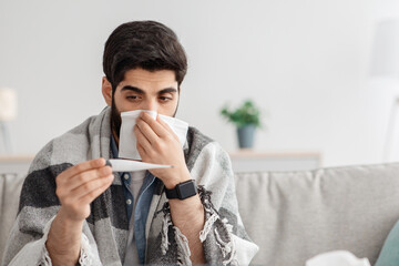 Cold, flu, coronavirus concept. Sick arab man sitting on couch and sneezing, measuring body...