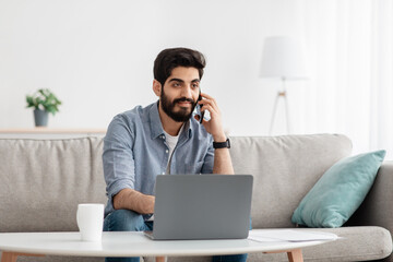 Always in touch. Arab freelancer man talking on cellphone using laptop working remotely, sitting on sofa, free space