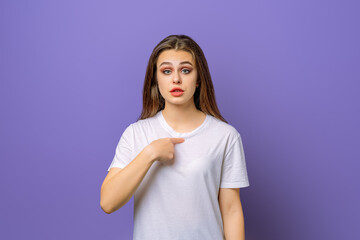 Fototapeta premium Surprise and puzzled young brunette woman pointing finger at herself, stands over purple background. Young emotional woman in blank white t shirt