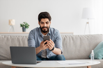 Business and telecommuting concept. Happy young arab man using smartphone and modern laptop computer, working at home