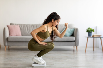 Excited young Indian woman in sportswear sitting on scales, making YES gesture, happy with result...