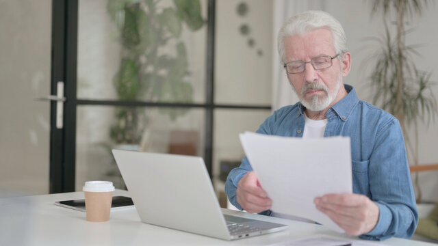 Senior Old Man Reading Documents while using Laptop in Office