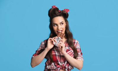 Sexy millennial pinup woman in retro outfit eating chocolate bar on blue studio background, panorama