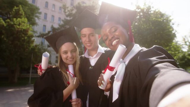 In the college garden multiracial graduates students young women and one guy using the camera to make a selfie video they are smiling large looking straight to the camera posing and holding diplomas