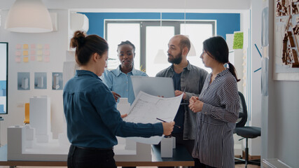 Multi ethnic group of architects working with blueprints and building model on table to design construction layout together. People doing teamwork on architectural urban project.