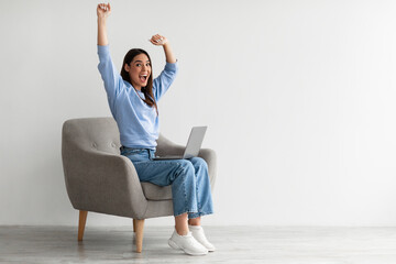 Full length of emotional woman with laptop raising hands, sitting in armchair, receiving great...