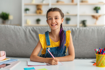 Positive girl sitting at table, writing and posing at camera