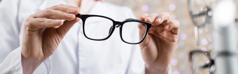 cropped view of oculist in white coat holding eyeglasses in optics salon, banner.