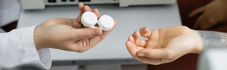 cropped view of ophthalmologist giving lens case to tattooed woman, banner.