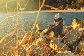 A woman in a jacket and a hat with a backpack sits on the rocks and watches the sea