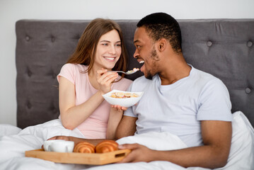 Obraz na płótnie Canvas Young Caucasian woman feeding her black husband with tasty cereal in bed at home