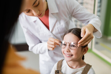 ophthalmologist trying eyeglasses on smiling girl near blurred mom in optics store. - obrazy, fototapety, plakaty