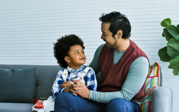 Handsome Asian single man taking care his African adopted child, smiling and holding hands with happiness, sitting on sofa in cozy living room at indoor home. Diversity, Kid and Education Concept.