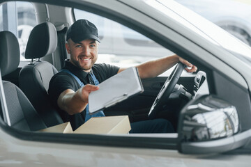 Cheerful worker. Delivery man in uniform is indoors with car and with order