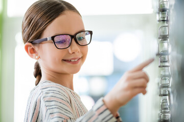 smiling girl in eyeglasses pointing with finger in optics store on blurred foreground.