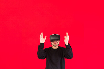 Man with cap dressed in black with Virtual Reality glasses smiling while touching something virtually, on red background, with copy space. Technology, virtual reality, computing and hobbies concept.