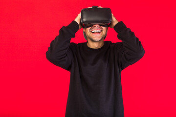 Man in cap dressed in black wearing Virtual Reality glasses, smiling, hands on head, surprised, on red background. Technology, VR, computing and hobbies concept.