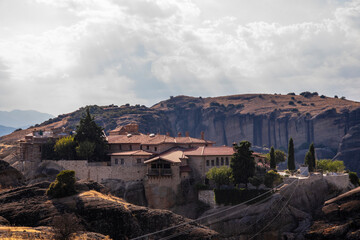 Fototapeta premium Beautiful scenic view of Orthodox Monastery of Áyios Ayía Triáda (Holy Trinity) on cliff, immense monolithic pillar, at the background of stone wall and rock formations of Meteora mountain, Greece.