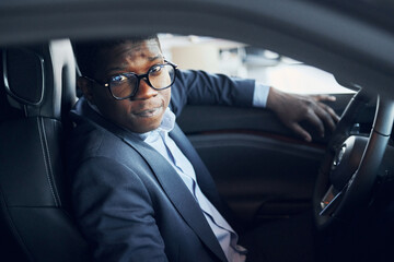 In glasses. Young african american businessman in black suit is in the automobile