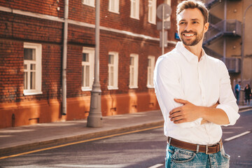 Fototapeta premium Portrait of handsome smiling stylish hipster lambersexual model.Modern man dressed in white shirt. Fashion male posing on the street background. Outdoors at sunset. Crossed arms