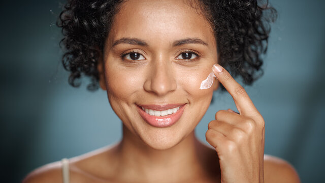 Female Beauty Portrait. Beautiful Black Woman With Afro Hair Touching Natural, Healthy Skin, Using Facial Moisturizer, Applying Face Cream. Wellness And Skincare Concept On Soft Isolated Background.