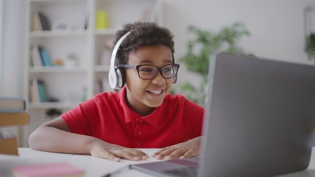 Joyful kid in headphones talking to friend online, using laptop application