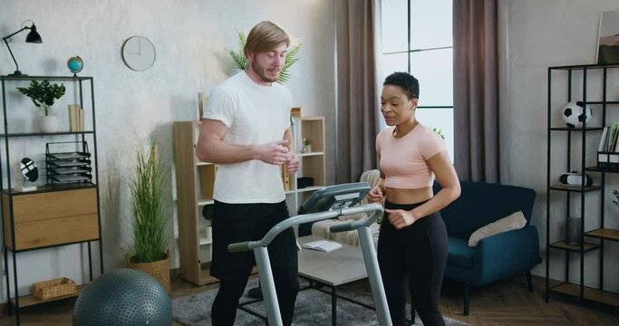Handsome sportive strong bearded guy in sportswear running on treadmill while his fit african american female friend mentoring him how to correct doing exercises