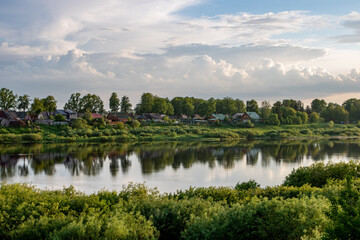 Fototapeta na wymiar small village rivers landscape in the evening with green trees, small houses on the coast, reflections on the water and clouthy sky
