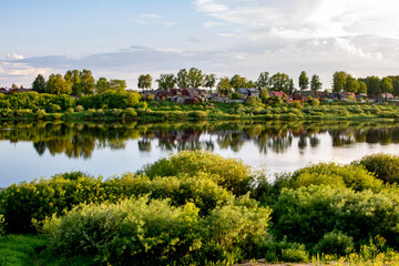 Fototapeta na wymiar village rivers landscape in the evening with green trees, small houses on the coast, reflections on the water and blue sky