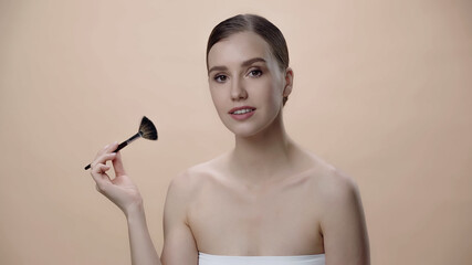 young woman with bare shoulders holding cosmetic brush isolated on beige.