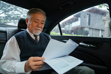 Confidence senior businessman in suit sitting on car backseat and reading business plan document while going to office. Elderly CEO working in automobile and looking at traffic out of the window