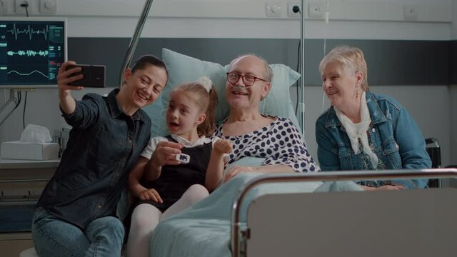 Family visiting retired man and taking selfies in hospital ward, mother and child using smartphone to have fun. Sick patient enjoying visit from niece and daughter with mobile phone at clinic.