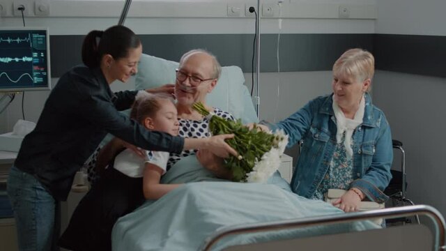 Close up of retired man receiving visit from niece and daughter in hospital ward. Little girl bringing flowers and visiting sick patient at medical clinic, giving support and comfort for recovery.