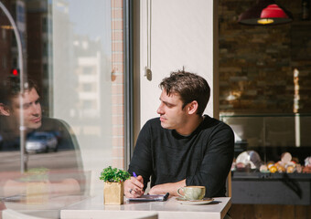 young man in a grey jacket writing something in note book while taking a coffee at the bar. Writing down his fresh ideas. Looking though the window. High quality photo