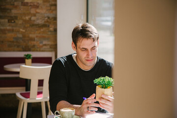  young man in a grey jacket holding a plant while taking a coffee at the bar while taking a coffee at the bar. Writing down his fresh ideas. High quality photo