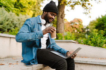 Black young man working with laptop while drinking coffee on bench