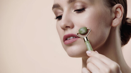 young woman using jade roller on face isolated on beige.