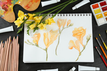 Painting of freesias in sketchbook, flowers and art supplies on black table, flat lay