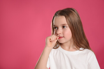 Cute little girl biting her nails on pink background, space for text