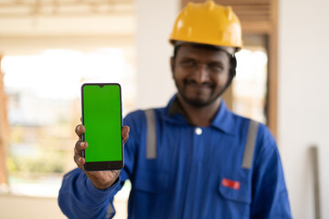 Focus on mobile pone, smiling plumber showing mobile phone with green screen mockup by lookling at camera - concept of booking of house repair service, app advertising and blue collar jobs