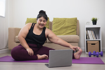 Young asian sport woman plus size sitting practicing yoga lesson. Stretching leg training workout at home for good health and body shape. Learning skill form video online class by laptop.