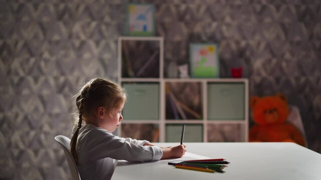 Upset little girl draws picture with crayon sitting at table