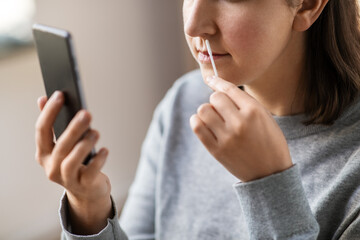 medicine, quarantine and pandemic concept - close up of woman with swab and smartphone taking sample from her nose and making nasal coronavirus self test at home - obrazy, fototapety, plakaty
