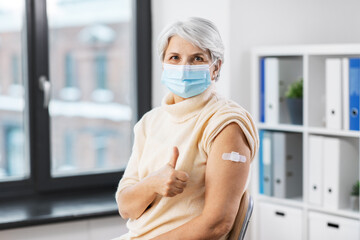 Fototapeta na wymiar medicine, health and vaccination concept - vaccinated senior woman in mask with medical patch on arm showing thumbs up gesture at hospital