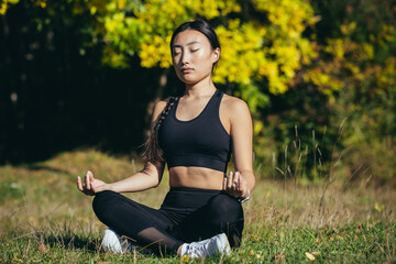 Young beautiful asian woman performs fitness yoga exercises, sitting in lotus position on the grass...