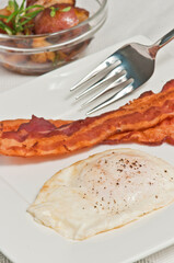 top view, close up of, a sunny side up, egg with pepper and two strips of broiled bacon with a fork and glass bowl of roasted potatoes