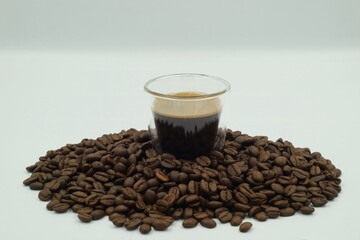 Espresso shot in a clear glass surrounded by coffee beans on a white screen.