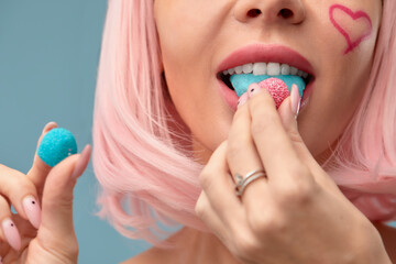 Close up of a flirtatious female model on blue background wearing a pink wig with a candy in mouth. Pretty glamorous woman pink hair charm sweets lifestyle enjoyment sugar