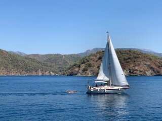 A yacht with a white sail on the open sea, against the backdrop of mountains. A sailboat floats in the ocean off the coast. A boat with a sail on the waves of a calm sea.